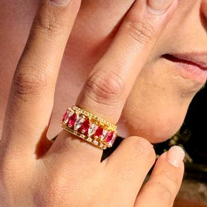 May include: A gold ring with alternating red and white marquise-cut gemstones. The ring is embellished with small, clear gemstones. The ring is worn on a finger.