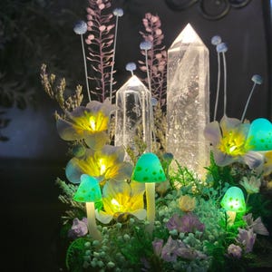 Pu&ograve; includere: Una composizione stravagante con funghi artificiali illuminati con cappucci verdi e steli bianchi, circondati da fiori gialli luminosi e punte di cristallo trasparente. La composizione include accenti floreali viola e rosa, creando un'estetica da favola.
