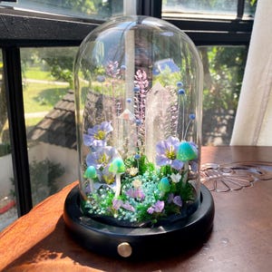 Pu&ograve; includere: Un terrario a campana di vetro con base nera, che mostra un giardino in miniatura. All'interno, ci sono fiori blu e viola, funghi verdi e un grande cristallo. La composizione include piccoli fiori bianchi e varie piante verdi.