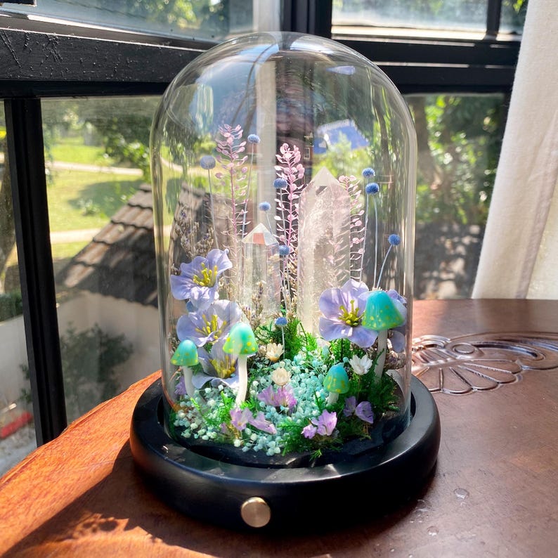 Pu&ograve; includere: Un terrario a campana di vetro con base nera, che mostra un giardino in miniatura. All'interno, ci sono fiori blu e viola, funghi verdi e un grande cristallo. La composizione include piccoli fiori bianchi e varie piante verdi.