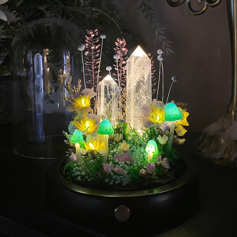 Pu&ograve; includere: Un terrario decorativo illuminato con cristalli di quarzo trasparente, funghi luminosi e fiori delicati. La composizione &egrave; ambientata in una base nera con un bordo dorato, creando un'esposizione stravagante e incantevole. I funghi e i fiori emettono una luce soffusa e calda.