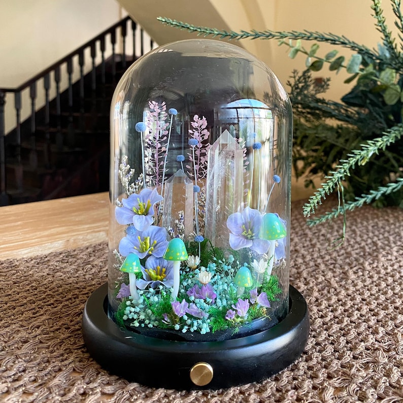 Pu&ograve; includere: Un terrario a campana di vetro con base in legno nero. All'interno, ci sono fiori blu, funghi verdi e punte di cristallo trasparente. La composizione &egrave; ambientata su uno sfondo di fiori secchi e vegetazione, creando un'esposizione stravagante.