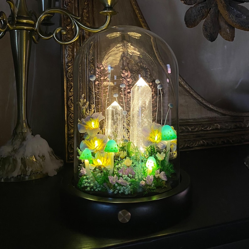 Pu&ograve; includere: Una cupola decorativa in vetro con cristalli, funghi e fiori illuminati. La base &egrave; marrone scuro con un bordo dorato. I cristalli e i funghi brillano di una luce soffusa, creando un'esposizione stravagante e incantevole.