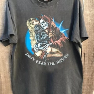 Op de afbeelding: Donkergrijs t-shirt met een afbeelding van de Grim Reaper die een vrouw omhelst. De Reaper draagt een rode cape met capuchon en de vrouw draagt een zwarte outfit. De tekst "DON'T FEAR THE REAPER" staat eronder.