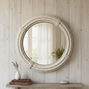 Può includere: Uno specchio rotondo con una spessa cornice in stile corda bianco sporco. Lo specchio è montato su una parete in legno chiaro. Un piccolo ripiano sottostante contiene libri, un vaso con lavanda e una pietra. Arredamento costiero.