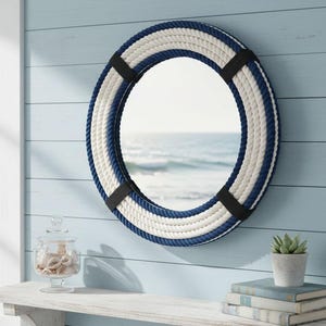 May include: Round nautical mirror with a rope design in white and navy blue. The mirror is mounted on a light blue wall. Black straps secure the rope. A coastal-themed decorative piece.