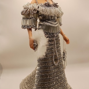 May include: A doll in a silver gown with matching accessories. The dress is made of small, metallic pieces, creating a textured effect. The doll has blonde hair and is wearing jewelry, a purse, and a feathered fan.