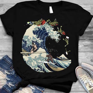 Peut inclure: T-shirt noir avec une parodie de "La Grande Vague de Kanagawa" de Hokusai. Le motif représente des personnages surfant et chevauchant la vague. La vague est en bleu et blanc, avec une montagne en arrière-plan. Design artistique et amusant.