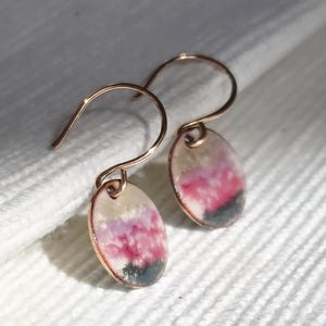 May include: A pair of handmade earrings with a copper-colored hook and an oval pendant. The pendant features a design with pink, white, and dark green colors. The earrings are displayed on a white, textured surface.