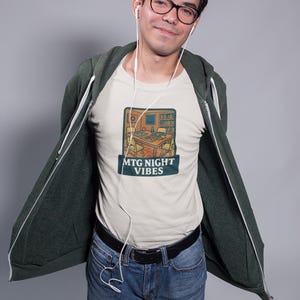 May include: A light-colored t-shirt with a graphic of a game room with the text "MTG NIGHT VIBES". The person is wearing a dark green zip-up hoodie, blue jeans, and glasses. White earbuds are visible.