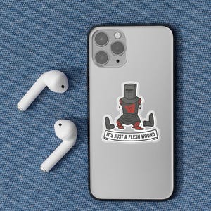 May include: A sticker on a phone depicts a cartoon knight with the text "IT'S JUST A FLESH WOUND." The knight is in black armor with red accents. White wireless earbuds are next to the phone on a blue denim surface.