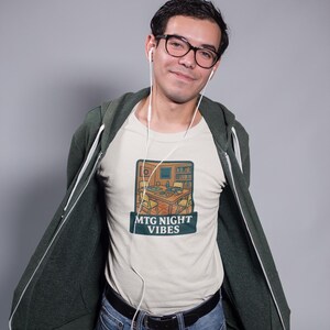 May include: A cream-colored t-shirt featuring a graphic illustration of a room with a table set for a card game, with the text "MTG NIGHT VIBES" printed below. The shirt is worn under an open dark green zip-up hoodie with white drawstrings.