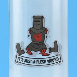 May include: A sticker featuring a black knight in armor with missing arms and legs, displaying a red emblem on the chest. Below the figure, a banner contains the text "IT'S JUST A FLESH WOUND" in black capital letters on a light background.