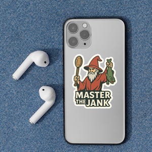 May include: A sticker on a phone features a cartoon wizard holding a football and a fish, with the text "MASTER THE JANK." Two white wireless earbuds are on the left.