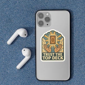 May include: A sticker on a phone with the text "TRUST THE TOP DECK" and a graphic of hands holding a card. Two white wireless earbuds are to the left of the phone. The background is a blue denim texture.