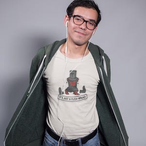 Könnte beinhalten: Ein cremefarbenes Kurzarm-T-Shirt mit einer Grafik eines dunkelgrauen Ritters ohne Gliedmaßen und dem Text "IT'S JUST A FLESH WOUND" auf einem Banner. Das T-Shirt wird unter einem offenen, dunkelgrünen Kapuzenpullover mit Reißverschluss getragen.