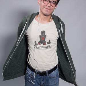 May include: A light-colored t-shirt with a graphic of a knight with the text "It's just a flesh wound." The knight is depicted in a seated position with visible injuries. The person is wearing a dark green zip-up hoodie and blue jeans.