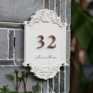 May include: Ornate white address plaque with the number 32 in bronze. The plaque has decorative scrollwork and the words "Sweet Home" in script. The plaque is mounted on a gray brick wall.