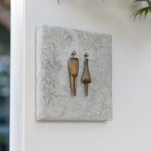 May include: A bronze metal restroom sign with abstract male and female figures. The sign is mounted on a square, grey, textured stone. The sign is fixed to a white wall.