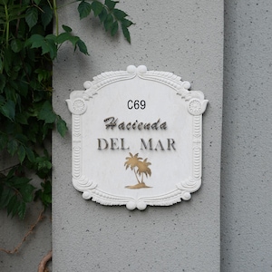 May include: White decorative sign with the text "C69 Hacienda DEL MAR" in black script and a gold palm tree illustration. The sign is mounted on a gray wall with green foliage in the upper left corner.