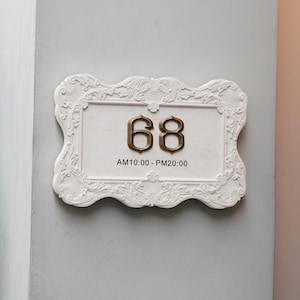 May include: A white, ornate plaque with the number "68" in gold-colored metal. The plaque has a decorative border and text that reads "AM10:00 - PM20:00". The plaque is mounted on a light gray wall.