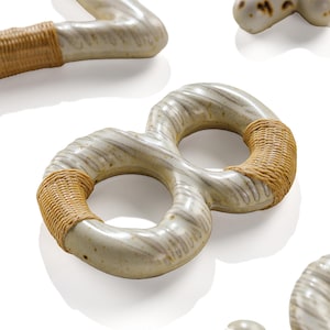 Pode incluir: Escultura de cerâmica branca suja em forma de símbolo de infinito, com detalhes em rattan tecido nas curvas. A cerâmica tem um padrão listrado texturizado. Outras esculturas semelhantes são visíveis no fundo.