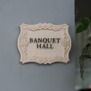 Könnte beinhalten: Cremefarbenes rechteckiges Schild mit dem schwarzen Schriftzug "BANQUET HALL". Das Schild hat einen dekorativen Rahmen mit floralen und scrollartigen Details. Es ist an einer hellblauen Wand befestigt.