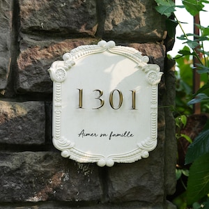 May include: A white, ornate address plaque with the number "1301" in gold lettering. The plaque has decorative scrollwork and the French phrase "Aimer sa famille" is written in script below the numbers. The plaque is mounted on a stone wall.