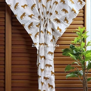 May include: White caftan dress featuring a repeating pattern of gold leopard illustrations. The dress has a V-neck and a drawstring waist. The text "Zodayhandmade" is printed on the fabric. The dress is hanging on a wooden door.