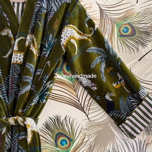 May include: Olive green velvet robe with a jungle animal print featuring leopards, zebras, and toucans. The robe has a striped trim on the sleeves and a matching belt. The background is a peacock feather pattern.