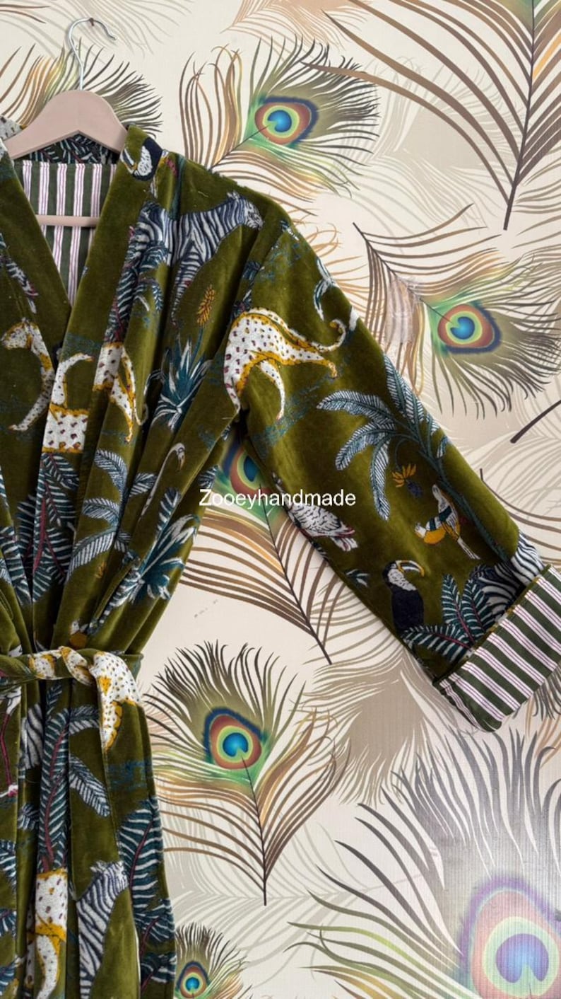 May include: Olive green velvet robe with a jungle animal print featuring leopards, zebras, and toucans. The robe has a striped trim on the sleeves and a matching belt. The background is a peacock feather pattern.