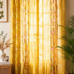 May include: A pair of sheer yellow curtains featuring a floral pattern and vertical striped panels. The curtains are hung from a gold-toned rod using fabric tabs. The fabric is translucent, allowing light to pass through, and displays the text ZoneyCurtains.