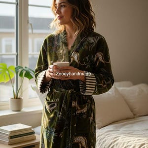 May include: A green velvet robe with a leopard and tropical leaf pattern. The robe has a shawl collar, a waist tie, and striped cuffs. The text "Zooeyhandmade" is visible on the robe. The person is holding a white mug.