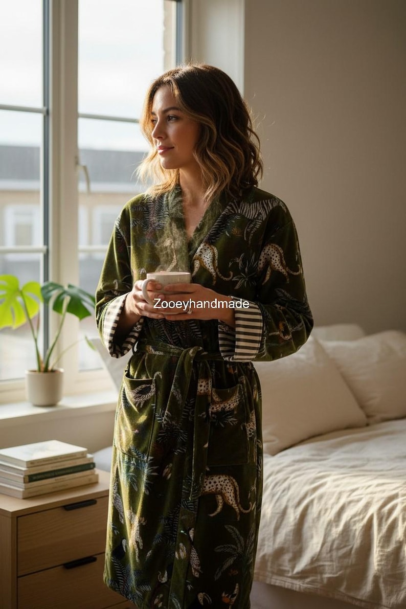 May include: A green velvet robe with a leopard and tropical leaf pattern. The robe has a shawl collar, a waist tie, and striped cuffs. The text "Zooeyhandmade" is visible on the robe. The person is holding a white mug.