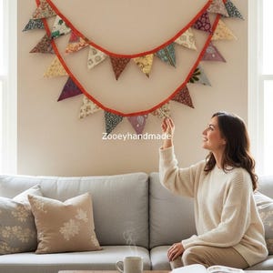 Può includere: Una ghirlanda di stoffa fatta a mano con bandierine triangolari in vari modelli e colori, infilate su un nastro rosso. La ghirlanda è appesa a un muro. Il testo "Zooeyhandmade" è visibile.