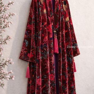 May include: A burgundy velvet robe with a floral pattern and pink tassels. The robe is hanging on a wooden hanger. The floral pattern includes red, pink, and blue flowers and green leaves. The robe has a purple lining.