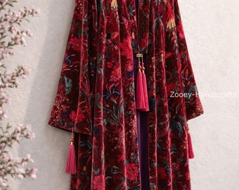 May include: A burgundy velvet robe with a floral pattern and pink tassels. The robe is hanging on a wooden hanger. The floral pattern includes red, pink, and blue flowers and green leaves. The robe has a purple lining.