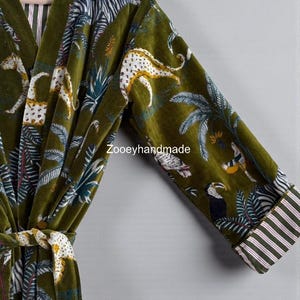 May include: Olive green velvet robe with a jungle animal print featuring zebras, leopards, and toucans. The robe has a striped trim on the sleeves and a matching belt. The robe is hanging on a wooden hanger.