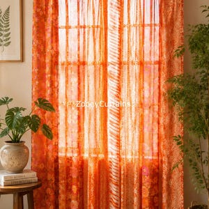 Pode incluir: Um par de cortinas laranja com uma mistura de padrões florais e listrados, penduradas numa vara dourada com presilhas. O tecido é semitransparente, permitindo a passagem de luz. O texto ZobeyCurtains é visível no centro das cortinas.