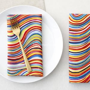 May include: A white plate with a gold fork and a colourful napkin with wavy stripes of red, yellow, blue, and black. Another napkin with the same design is to the right.