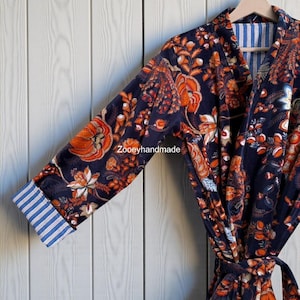 Könnte beinhalten: Ein marineblauer Bademantel mit orangefarbenem Blumenmuster und blau-weiß gestreiften Manschetten. Der Bademantel hängt an einem Holzbügel vor einem weißen Holzhintergrund. Der Text "Zooeyhandmade" ist sichtbar.