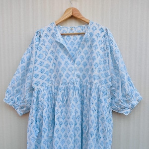 Può includere: Un abito midi bianco con un motivo floreale azzurro stampato a blocchi. L'abito presenta uno scollo a V con sottili laccetti, maniche a tre quarti a sbuffo con polsini elasticizzati e una vita arricciata che scende in una gonna.