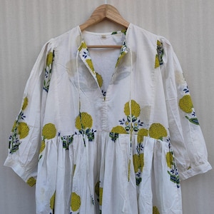 May include: White cotton dress with a floral pattern in yellow and blue. The dress has a V-neckline with a drawstring, three-quarter sleeves, and a tiered skirt. The dress is hanging on a wooden hanger.