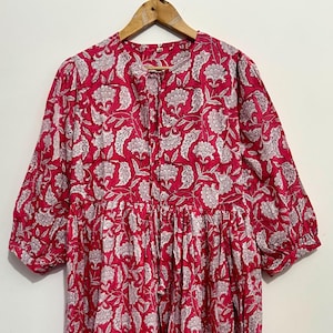 May include: A red dress with a white floral pattern. The dress has three-quarter sleeves and a tiered skirt. It is hanging on a wooden hanger.