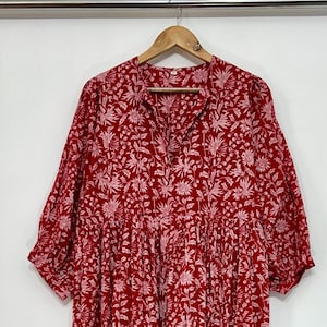 May include: A red dress with a white floral design. The dress features a V-neck, short sleeves, and a tiered skirt. It is displayed on a wooden hanger against a white backdrop.