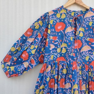 May include: A blue floral print dress with long sleeves. The dress features a vibrant pattern of red, yellow, and pink flowers and leaves. The dress is hanging on a wooden hanger.