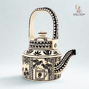 May include: Cream-colored ceramic teapot with black geometric and floral patterns. The teapot features a curved handle, a spout, and a lid. The design includes images of birds and a cup. The brand logo is visible in the upper right corner.