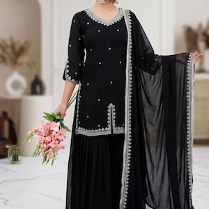 May include: Black embroidered salwar kameez suit with a matching dupatta. The suit features a V-neckline and intricate silver embroidery along the edges and scattered across the fabric. The model is holding a bouquet of pink flowers.
