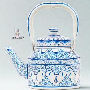 Può includere: Una teiera in ceramica bianca e blu con un beccuccio curvo e un'ampia maniglia. La teiera è decorata con intricati motivi e disegni floreali blu. Il coperchio ha un piccolo pomello. Il testo "LOOM & BLOOM" è visibile.