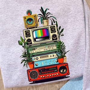 Puede incluir: Sudadera gris claro con un diseño gráfico de aparatos electrónicos vintage apilados, incluyendo una radio, un reproductor de cintas y un televisor. El diseño incorpora plantas en macetas y altavoces, creando una estética retro. La sudadera se combina con vaqueros azules y un sombrero.